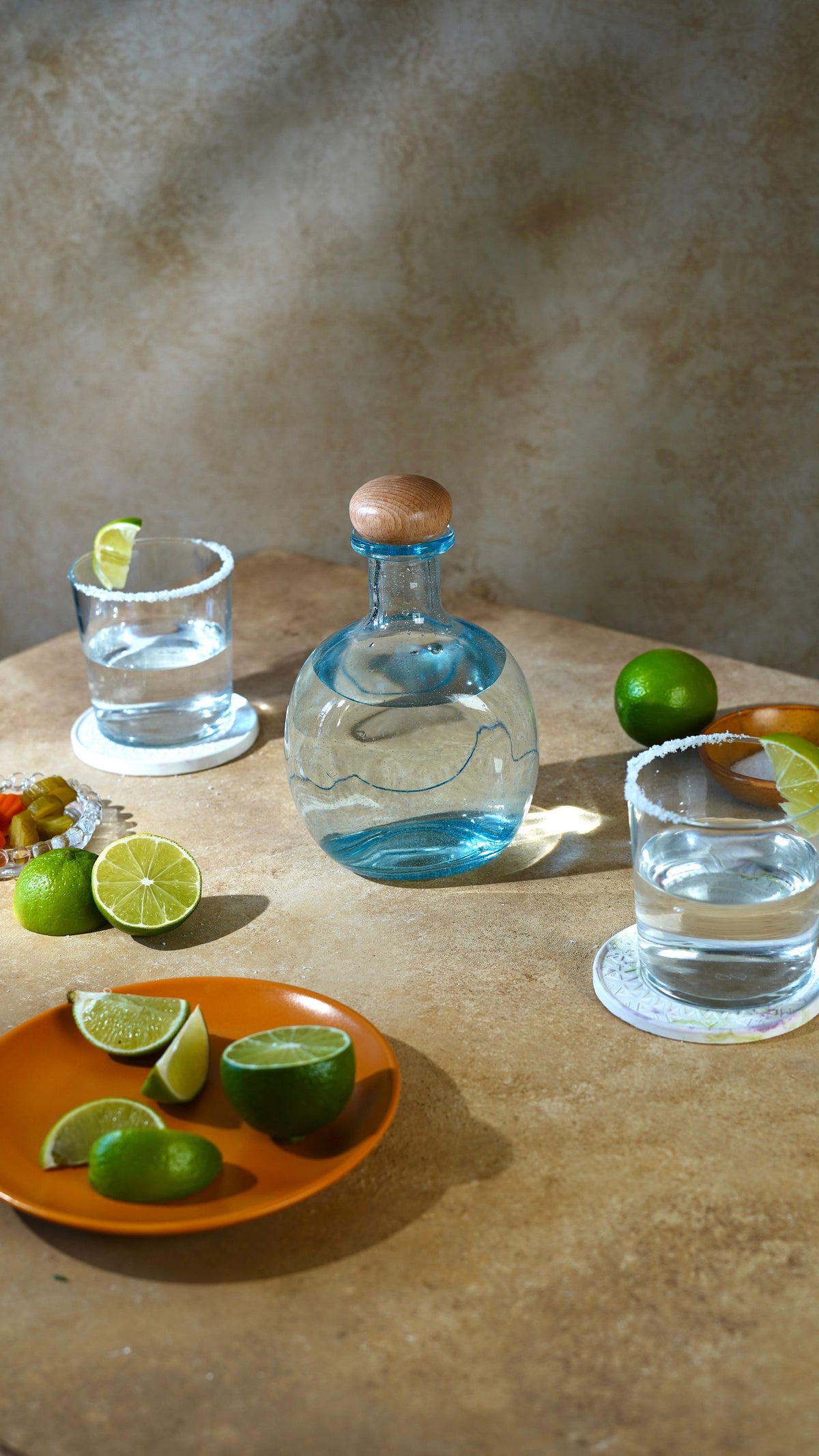 A hand-blown glass tequila decanter with salt-rimmed glasses and fresh limes on a stone table