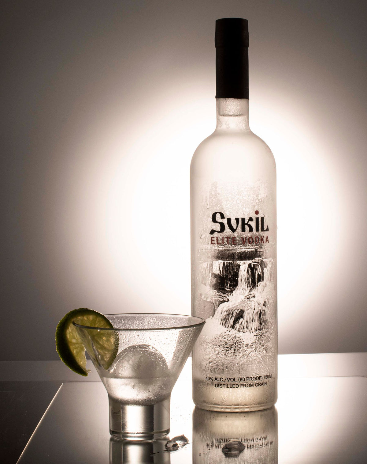 A clear vodka bottle beside a martini glass and lime under warm lighting
