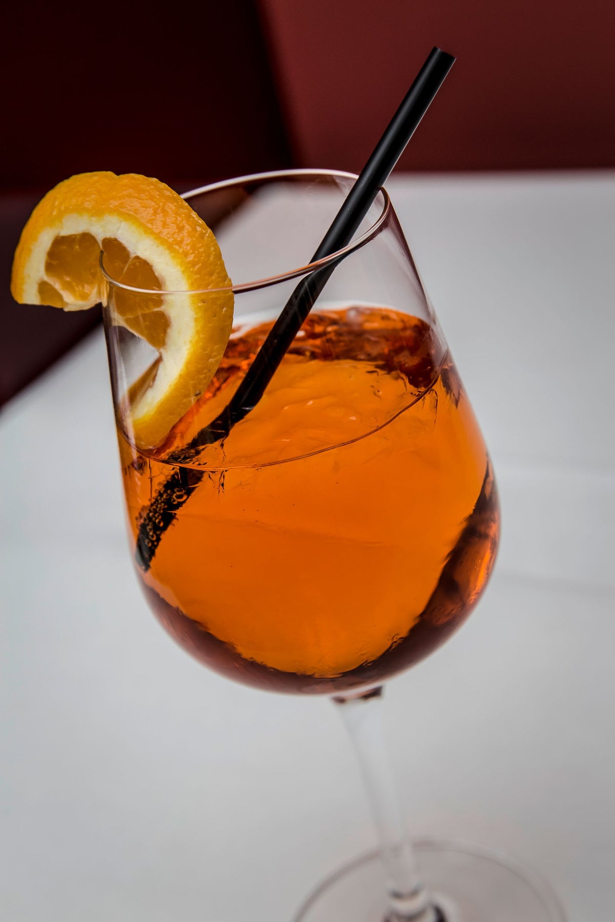 A bright orange spritz in a wine glass with ice, an orange slice on the rim and a black straw