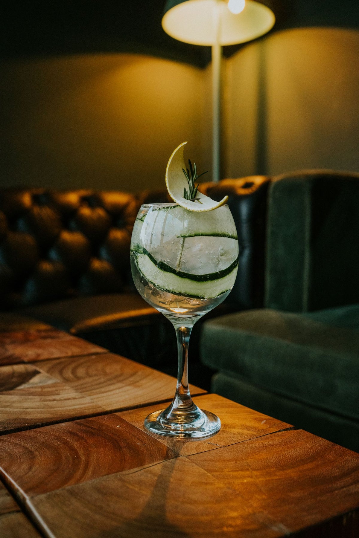A gin and tonic garnished with cucumber and rosemary on a wooden table in a dim bar