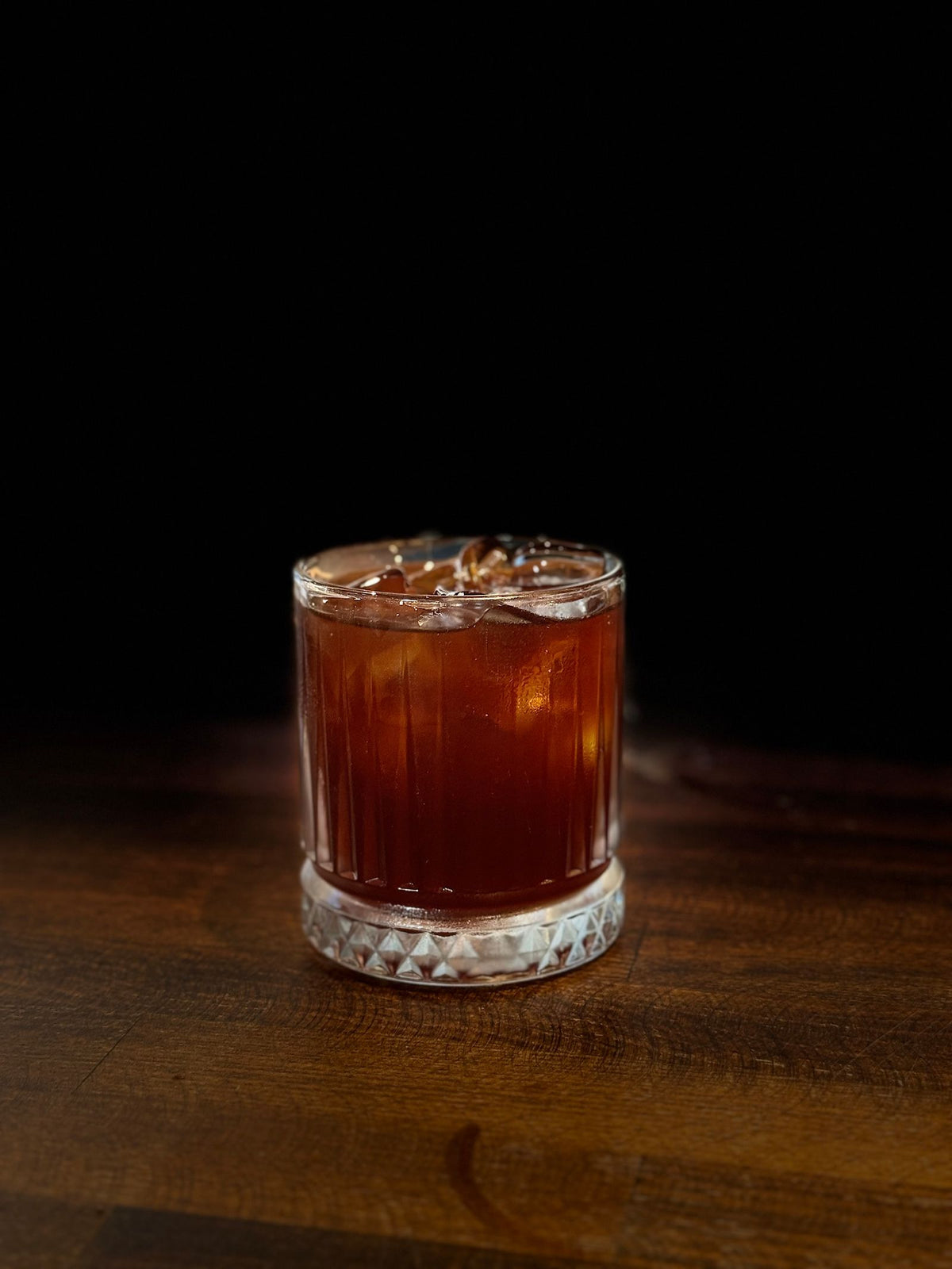 A glass of dark amber rum over ice on a wooden bar top