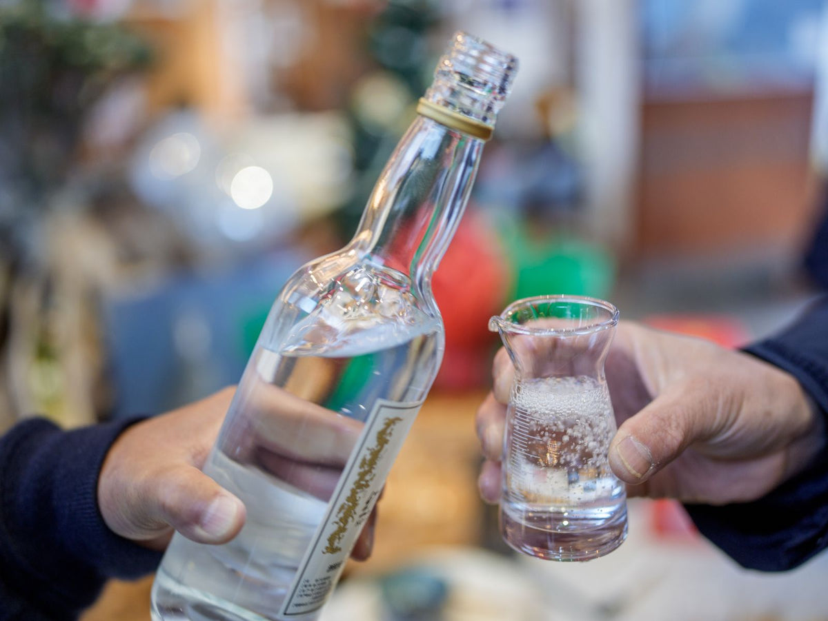 Hands pouring clear sake from an unlabelled bottle into a small glass carafe