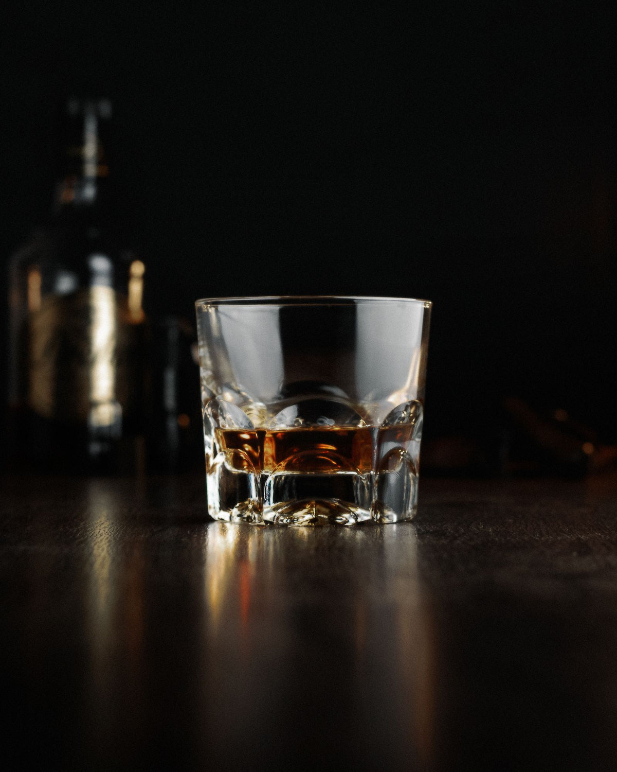 A glass of amber single malt whisky on a dark bar top, bottle out of focus behind it