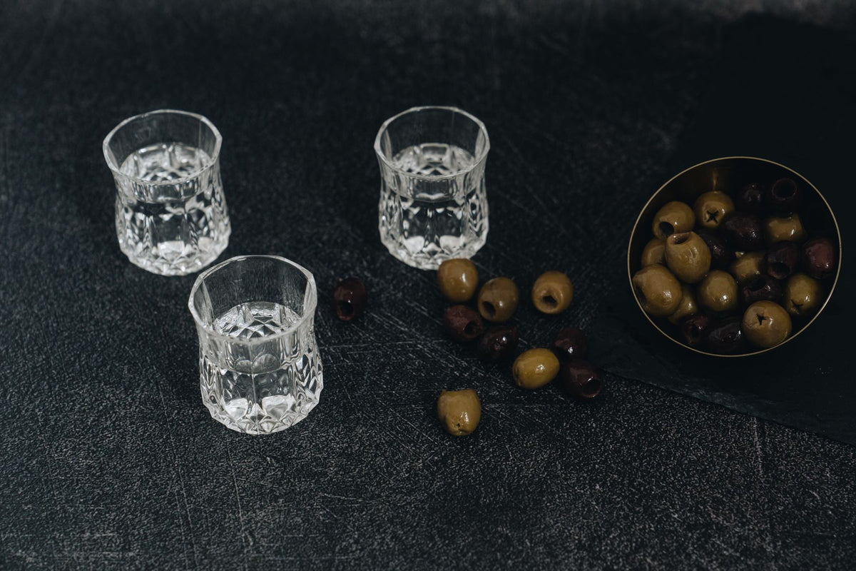 Crystal glasses of vodka beside a bowl of olives on a dark surface